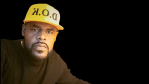 A confident Black man wearing a yellow D.O.K. hat and a black sweater, looking directly at the camera against a solid black background.