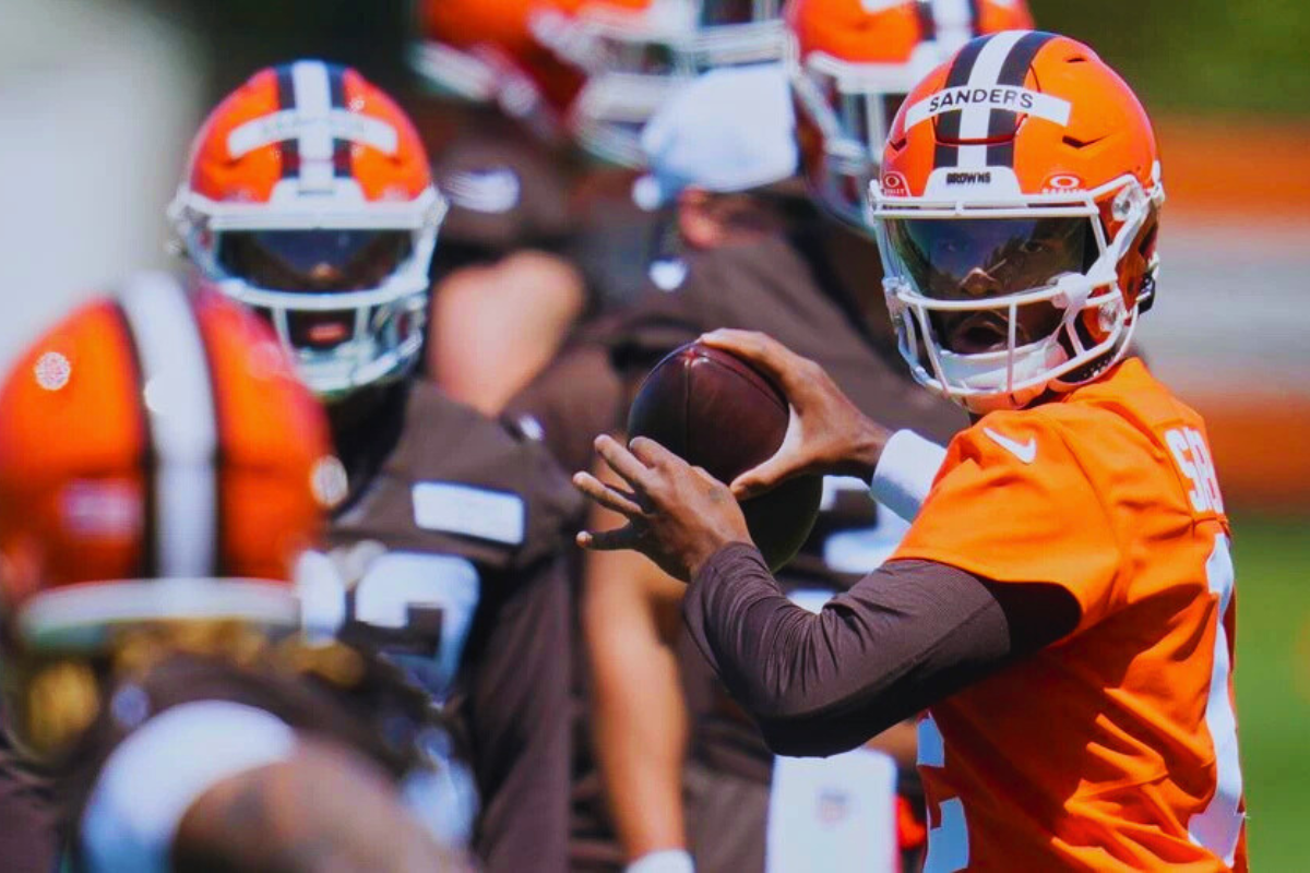 Shedeur Sanders in Cleveland Browns practice uniform, holding a football mid-throw with focused determination during training camp.
