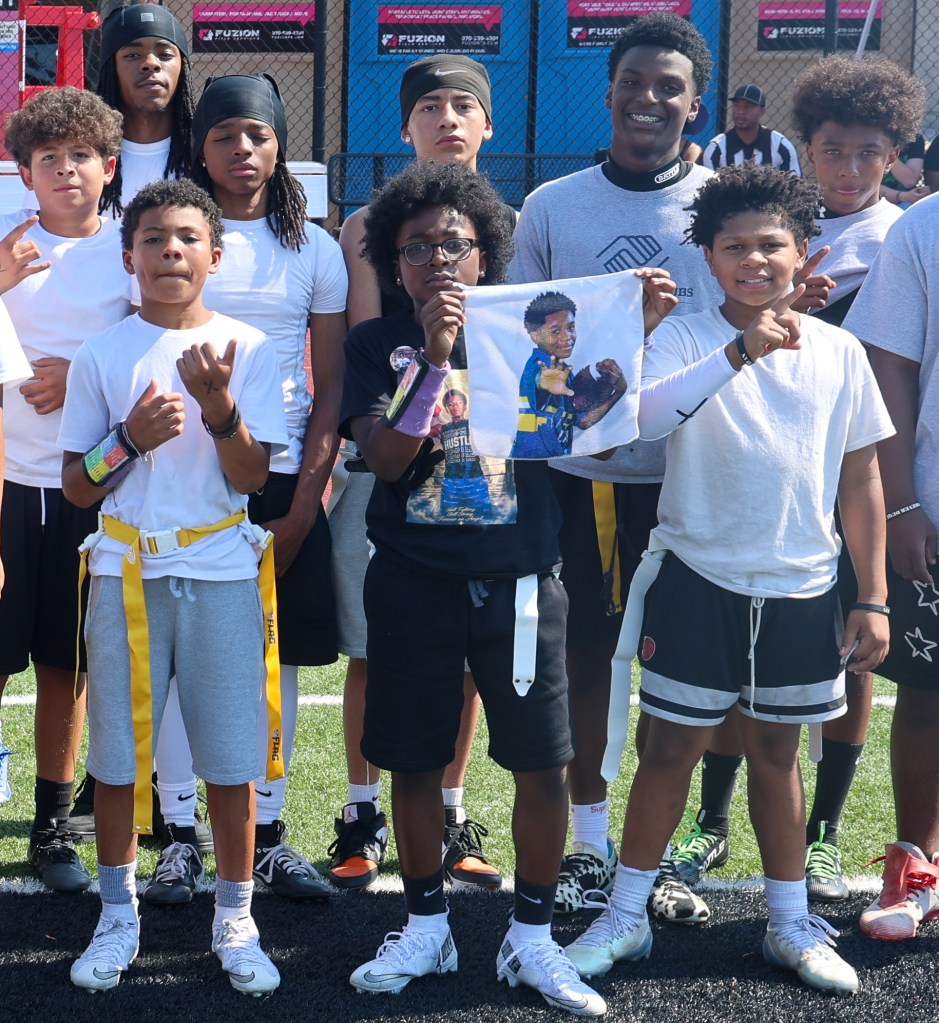 A group of youth flag football players pose together on the field while one participant holds a memorial towel featuring De'Quan during the Change for De'Quan community event in Denver, Colorado.