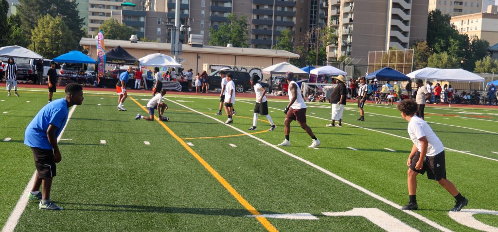 Youth football players line up before the snap while families, vendors, and community members gather around the field during the Change for De'Quan event.