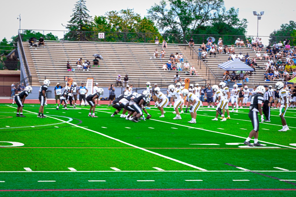 High school football teams line up at the line of scrimmage during a Saturday game, with players in black uniforms facing players in white uniforms as spectators watch from the stadium bleachers.
