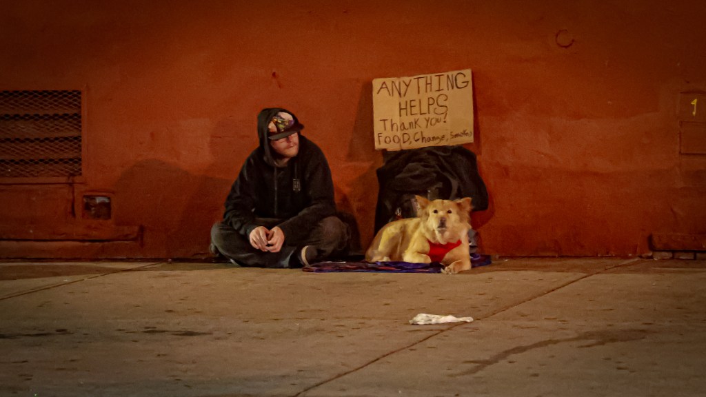 Two people sitting against an orange wall on a city sidewalk with a dog and a cardboard sign reading “Anything Helps.”