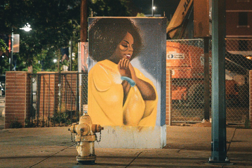 Colorful portrait artwork of a smiling woman in yellow displayed along a city sidewalk at night.