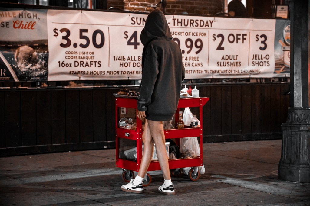 Hooded street food vendor standing beside a red food cart outside a bar at night.