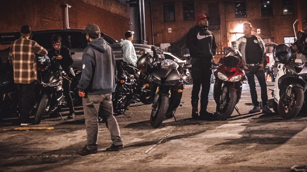 People gathered around motorcycles in a city parking area under streetlights at night.