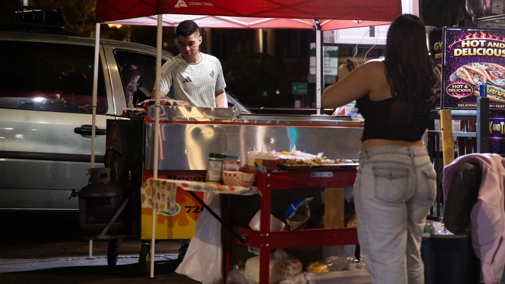 Street food vendor preparing food beneath a red canopy for a customer at night.