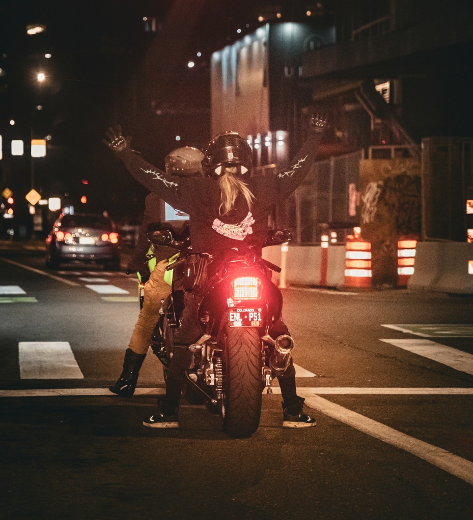 Motorcycle passenger raising both arms while stopped on a city street at night.