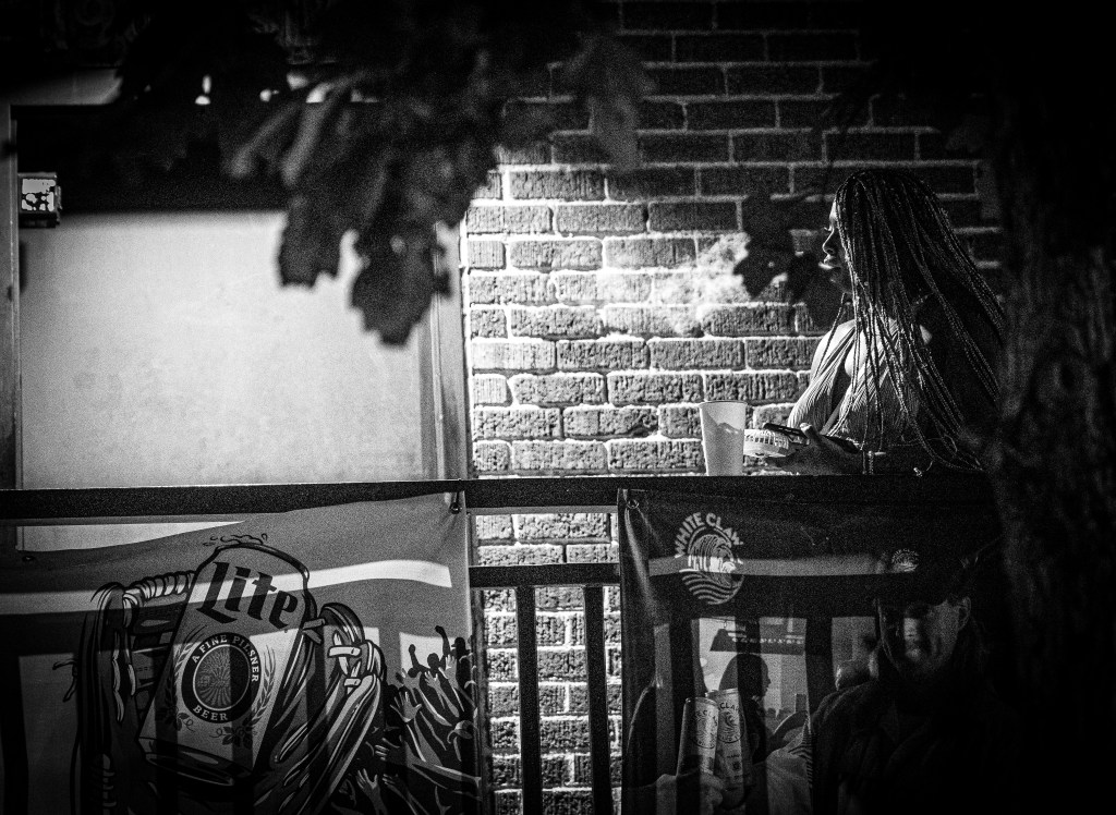 Black-and-white photograph of a woman with long braids standing near a brick wall at night.